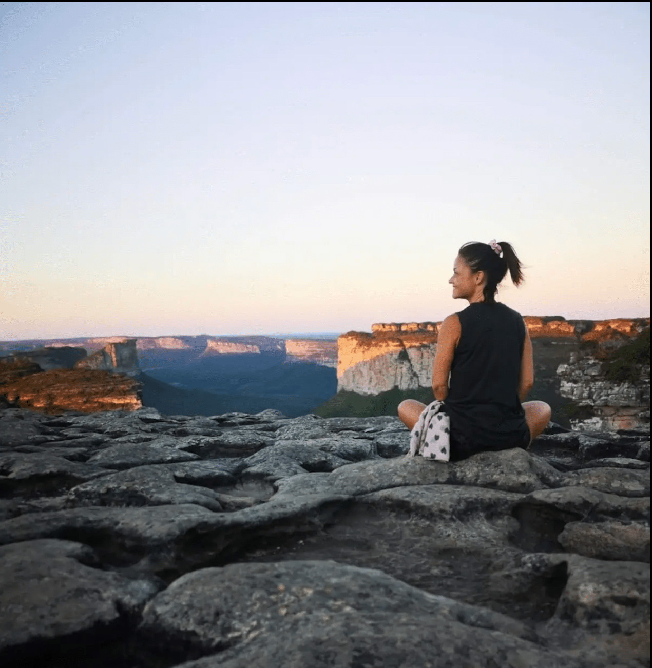 A woman sits cross-legged on rocky terrain at sunset, a peaceful expression on her face as she practices mindfulness with breathwork. Beside her rests a bag, symbolizing the calm found through the Intuitional 1-1 Session.
